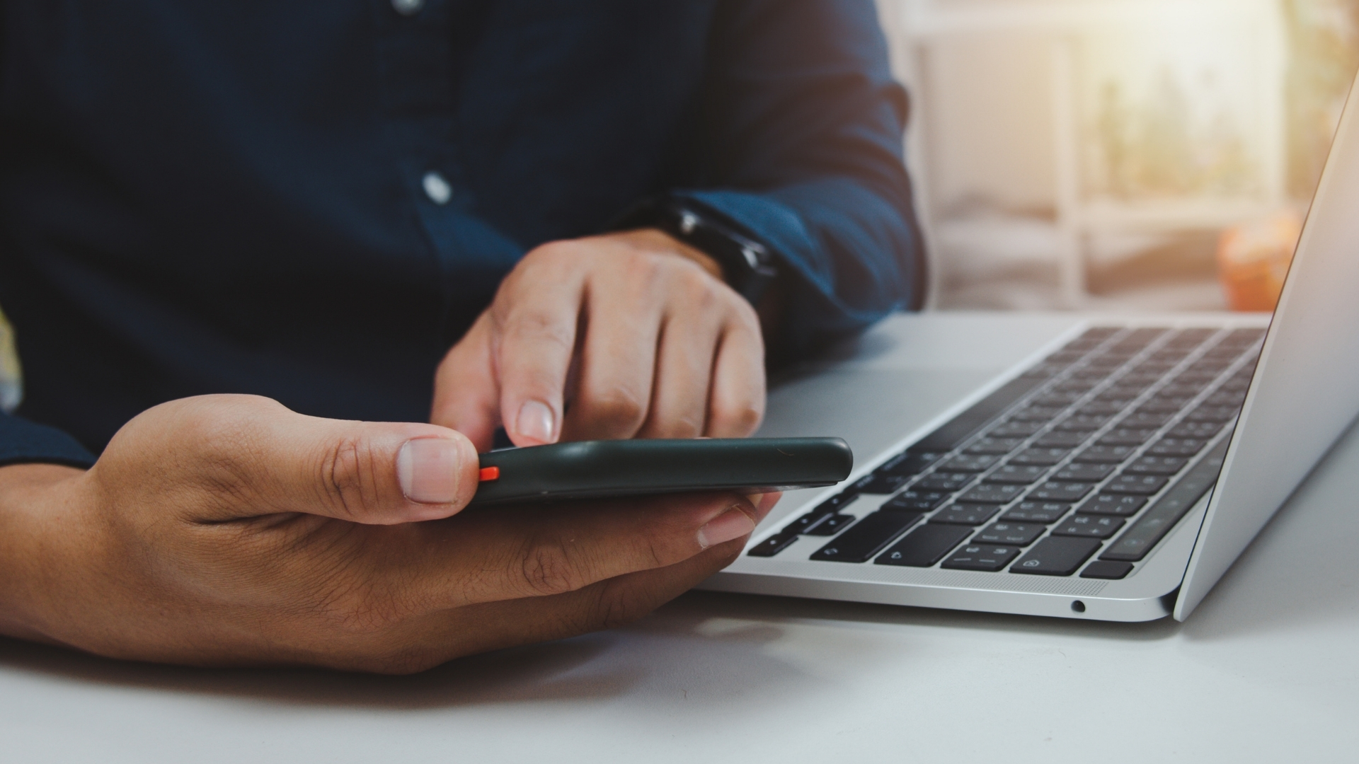 Homem de camisa azul e relógio usando um iPhone ao lado de um computador MacBook, ilustrando o tema da presença digital para pequenos negócios.